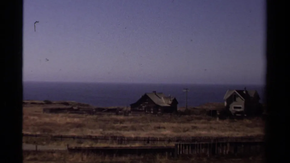 1980 8mm coastal view near Mendocino, showing bluff-side houses, dry grass, fence posts, a utility pole, and the ocean horizon.