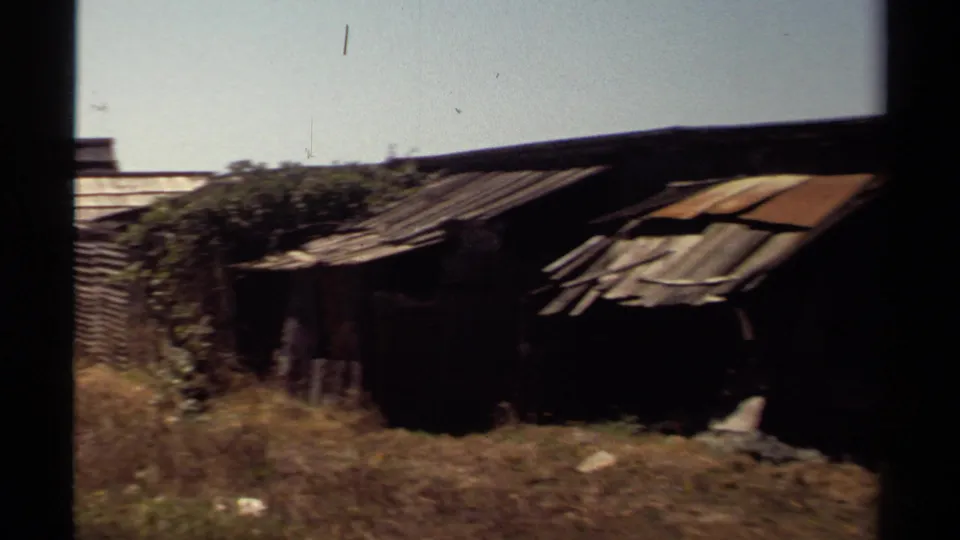 1980 Mendocino home-movie view of a low shed with patched roof panels, brush, fencing, and dark film-edge framing.