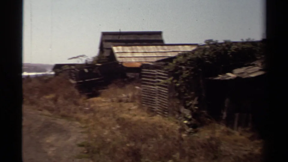 1980 Mendocino frame showing weathered sheds, rough fencing, a dirt path, dry grass, and a small glimpse of coastal water.
