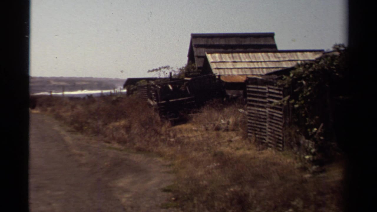 1980 Mendocino home-movie frame showing a dirt path, rustic wood buildings, dry grass, and distant sea or bay water.