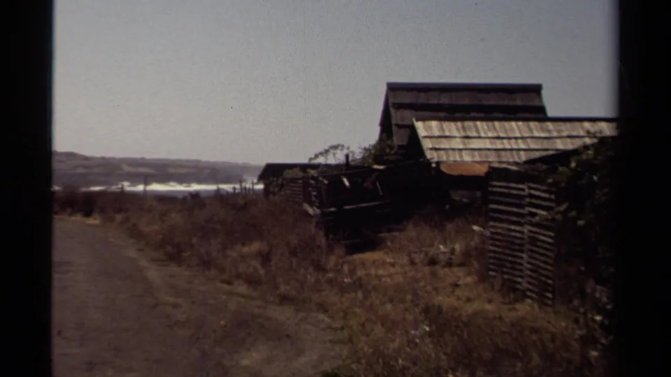 1980 Mendocino frame with rustic wooden outbuildings, rough fencing, dry vegetation, and distant coastal water.