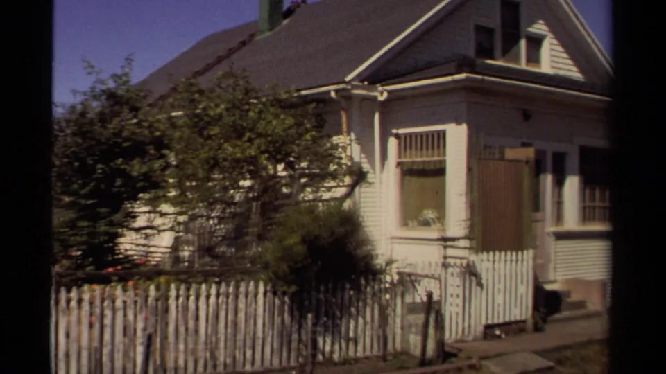 1980 Mendocino home-movie view of a light clapboard house, picket fence, shrubs, windows, and roofline.