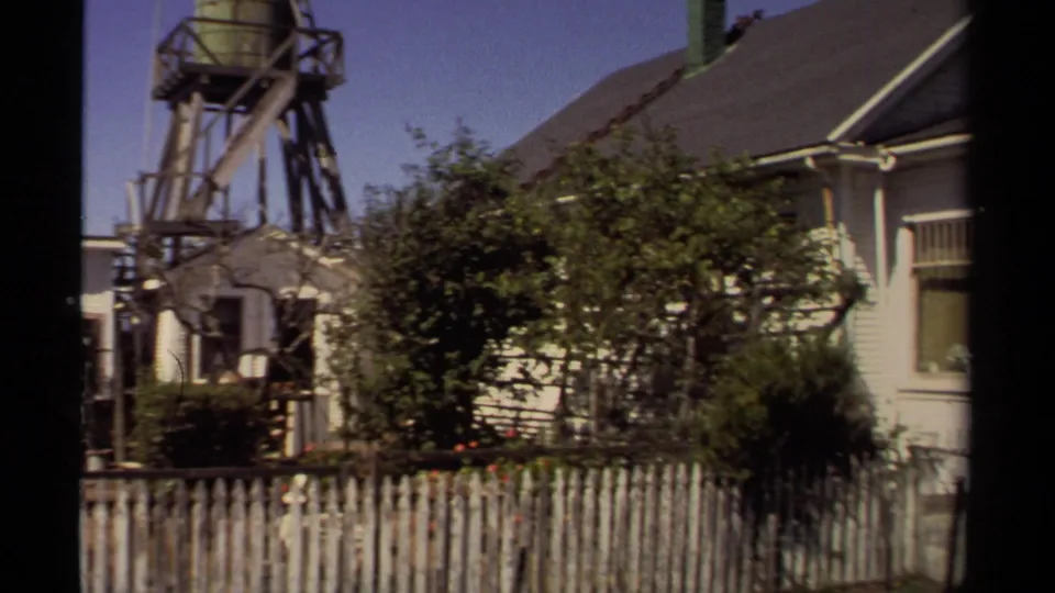 1980 Mendocino frame showing a house exterior, white picket fence, shrubs, and a small water tower structure behind the roofline.