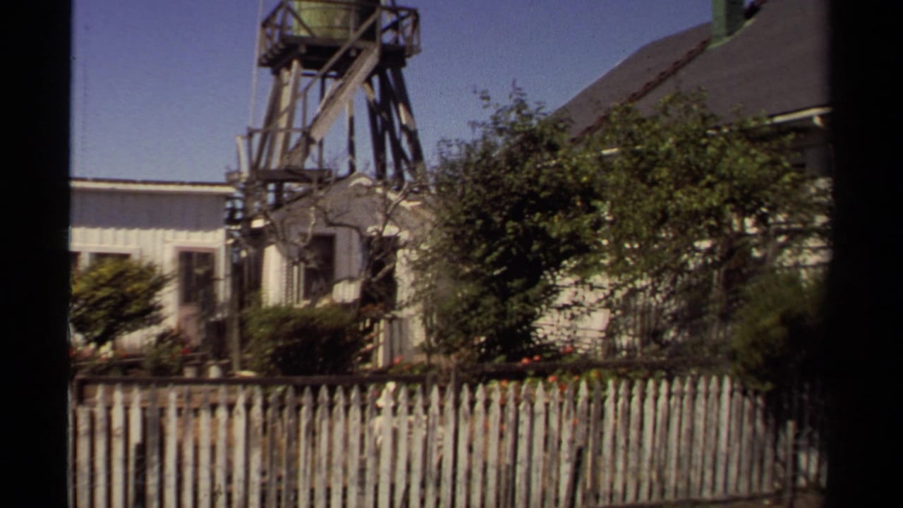1980 Mendocino home-movie frame showing clapboard buildings, garden shrubs, picket fencing, and a wooden water tank structure.