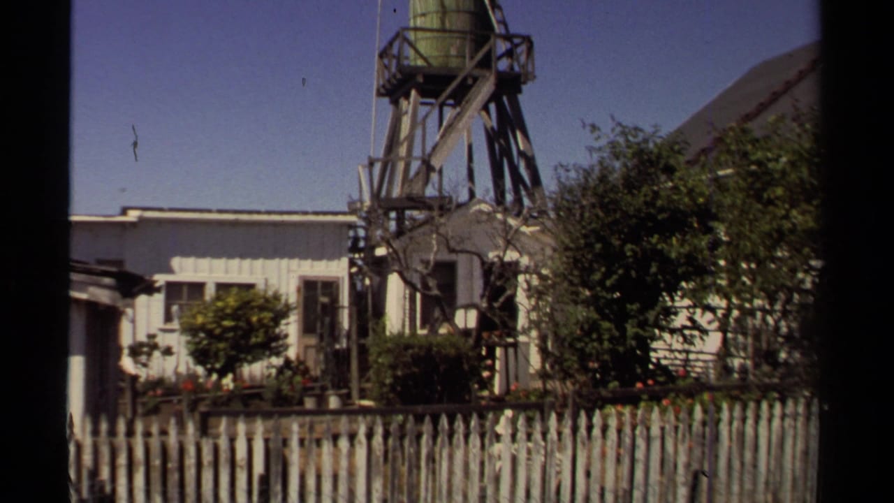 Raised water tank and residential buildings partly obscured by trees and a fence.