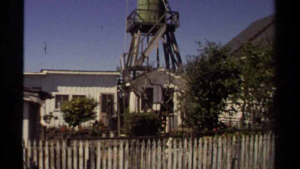 Mendocino neighborhood view with a green water tank on timber supports above clapboard buildings and dense garden plantings.