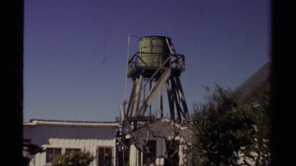 Handheld 8mm frame from Mendocino showing the elevated water tank, nearby rooflines, shrubs, and a white building below.