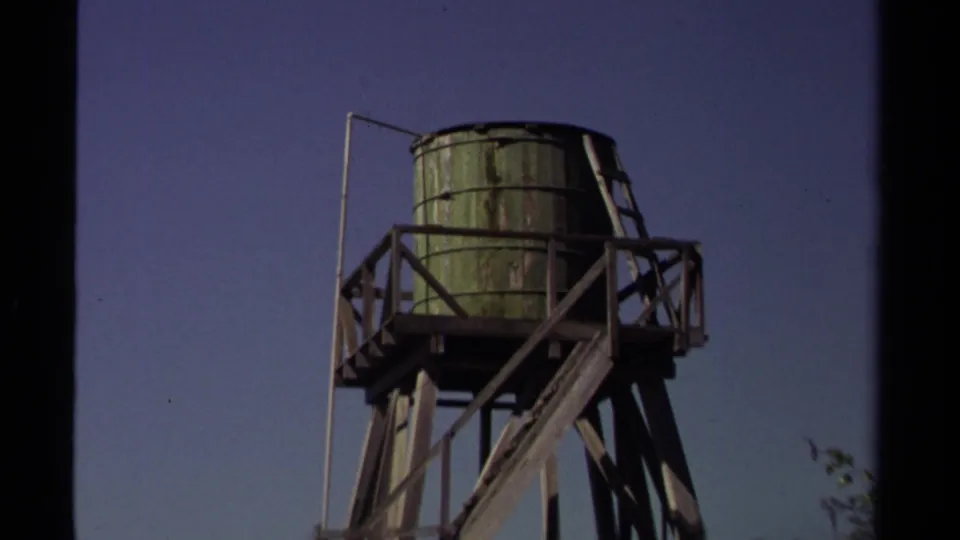 Tight 1980 Mendocino view of a raised green water tank, wooden railings, diagonal braces, and open blue sky.