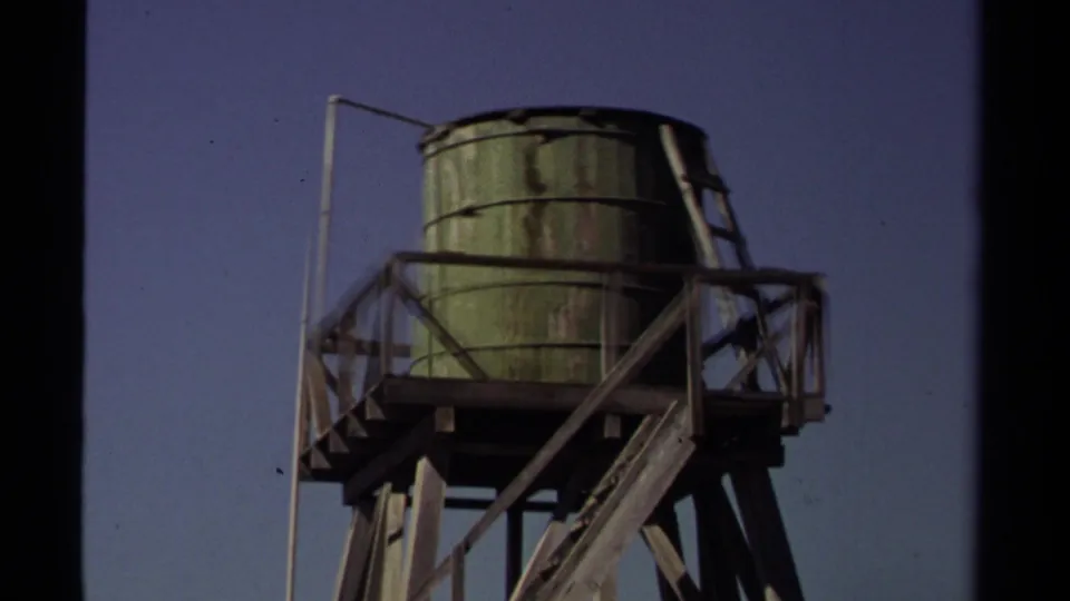 1980 Mendocino 8mm frame showing a weathered green water tank elevated on a wooden support structure under clear sky.