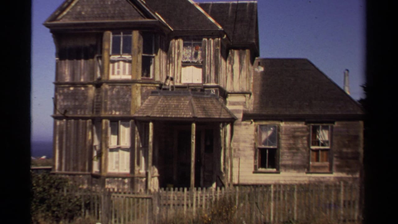 1980 Mendocino exterior view of a weathered two-story wooden house with peeling siding, porch, windows, and a simple fence.