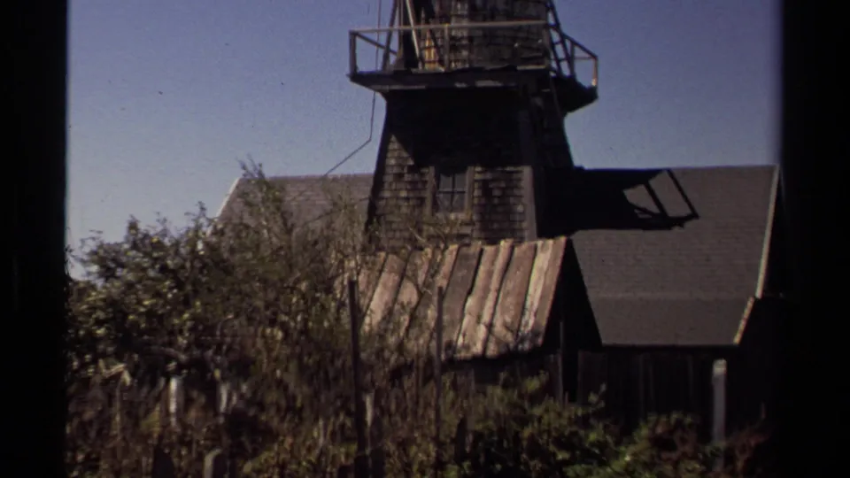 Mendocino exterior detail showing a weathered raised wooden section, railings, shrubs, roof surfaces, and clear sky in 1980 8mm footage.