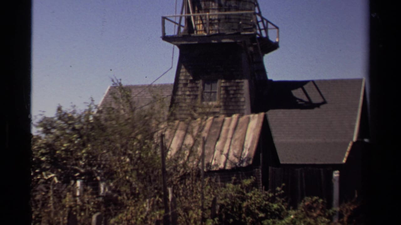 1980 Mendocino home-movie frame showing a weathered wooden building with a tower-like upper section, shrubs, and clear sky.