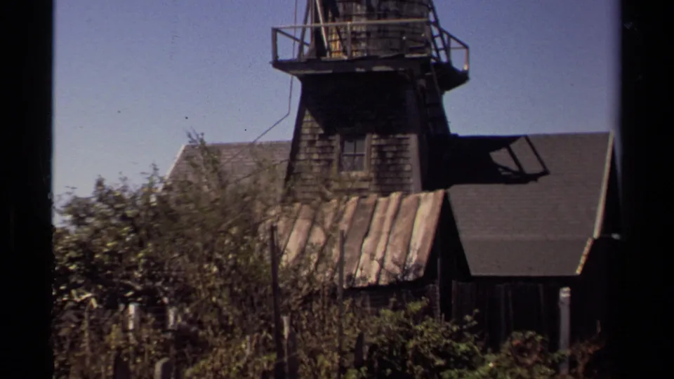 1980 Mendocino home-movie frame showing a weathered wooden building with a tower-like upper section, shrubs, and clear sky.