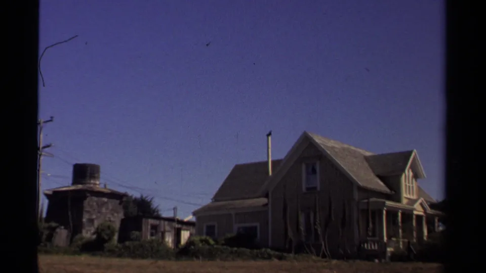 1980 Mendocino exterior view of a wooden house beside a small outbuilding and open yard beneath a clear blue sky.