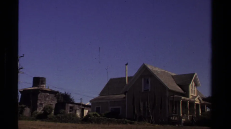 1980 Mendocino home-movie view of a wooden house and adjacent outbuilding across a quiet yard, framed by dark film edges.