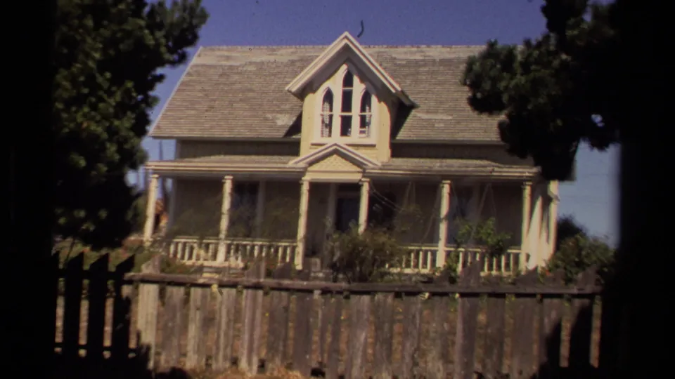 1980 Mendocino frame showing a two-story farmhouse behind a weathered wooden fence, with trees and shrubs around the porch.