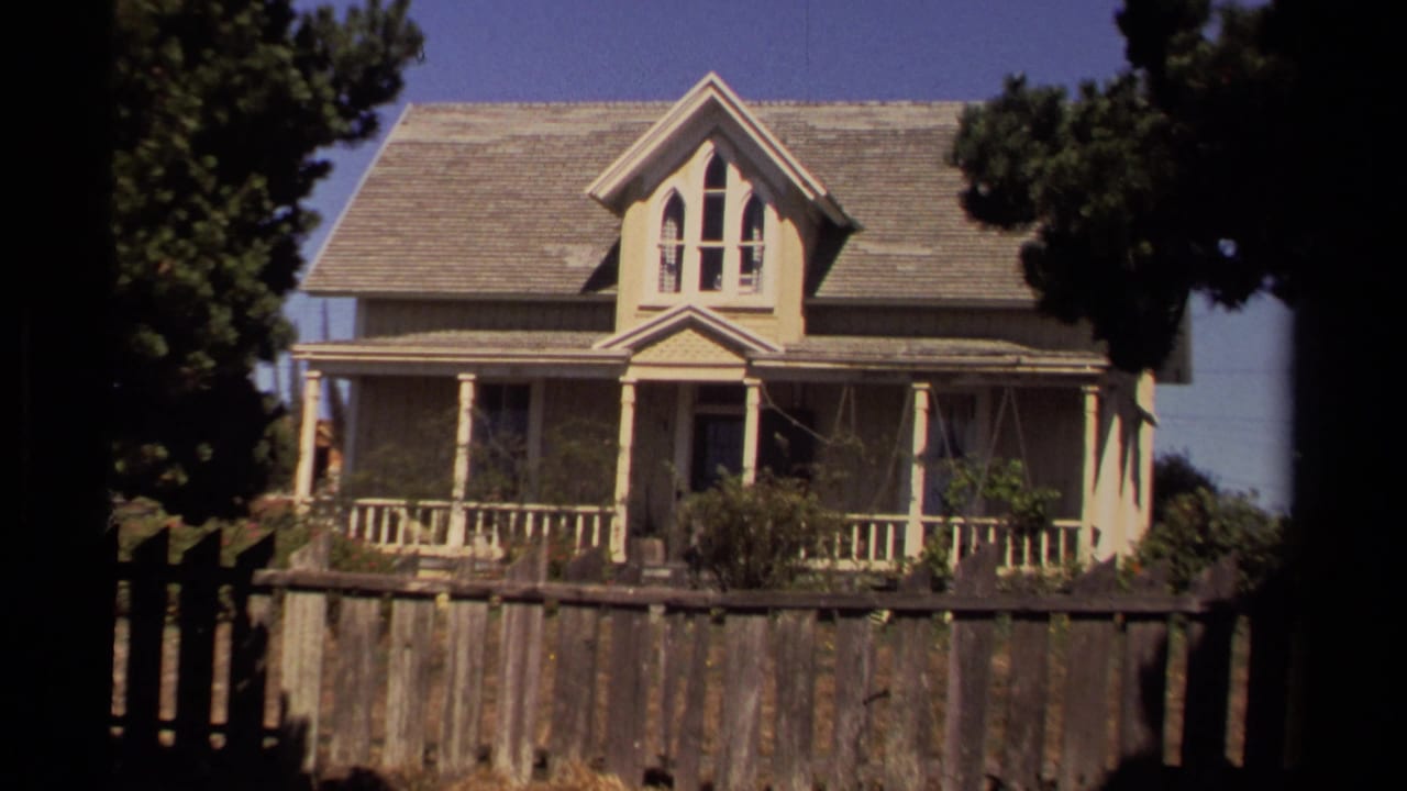Gabled wooden house with porch behind a weathered fence and trees.