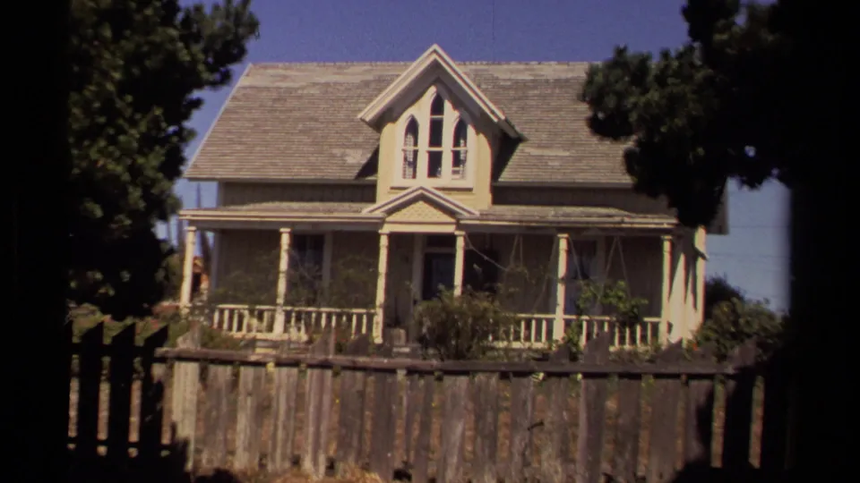 1980 Mendocino home-movie frame showing an older gabled wooden house with a front porch, garden, fence, and trees.