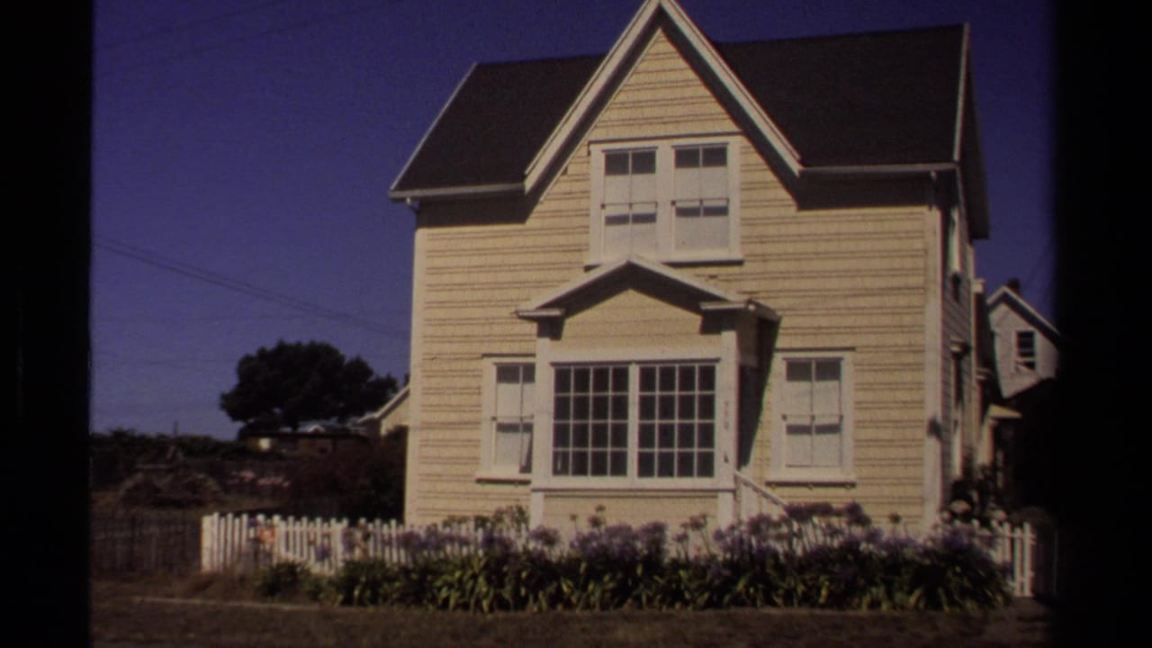 1980 Mendocino frame of a pale clapboard house with a steep gable, shuttered windows, white picket fence, and flower bed.