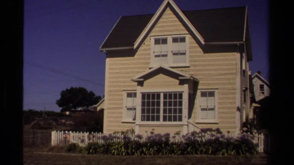 1980 Mendocino frame of a pale clapboard house with a steep gable, shuttered windows, white picket fence, and flower bed.