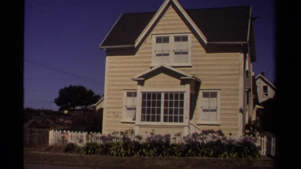 Closer 1980 Mendocino view of a pale clapboard house with a front window, shutters, flower bed, and low white fence.