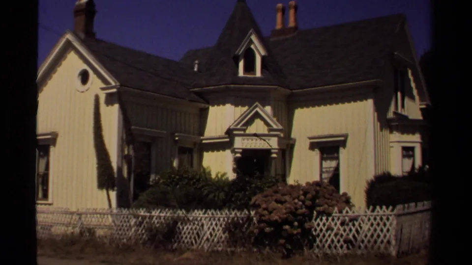 1980 Mendocino frame showing a yellow Victorian-style house with a dark roof, decorative entry, shrubs, and a white lattice fence.
