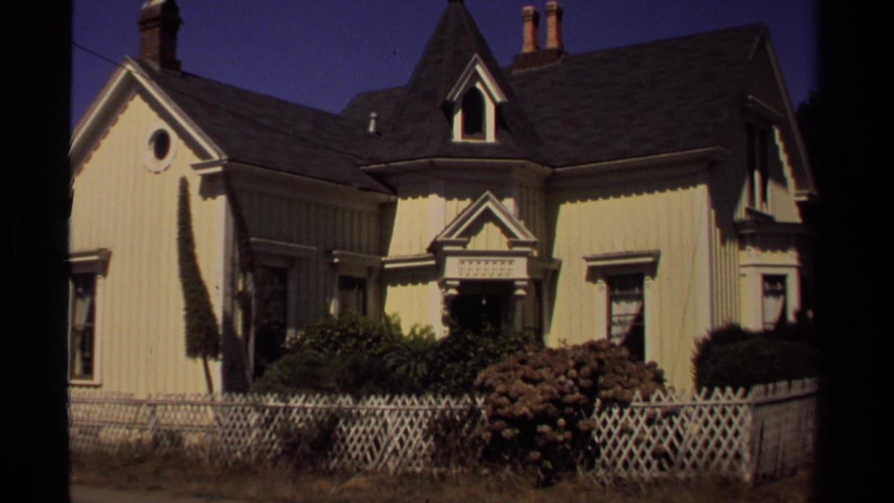 1980 Mendocino home-movie frame showing a pale Victorian-style house with gables, trim, shrubs, and a white lattice fence.