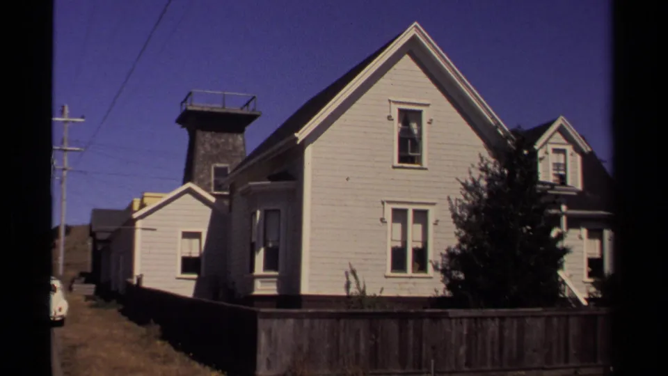 1980 Mendocino frame of a light residential building beside a narrow street, with a wooden fence, tree, and gabled rooflines.