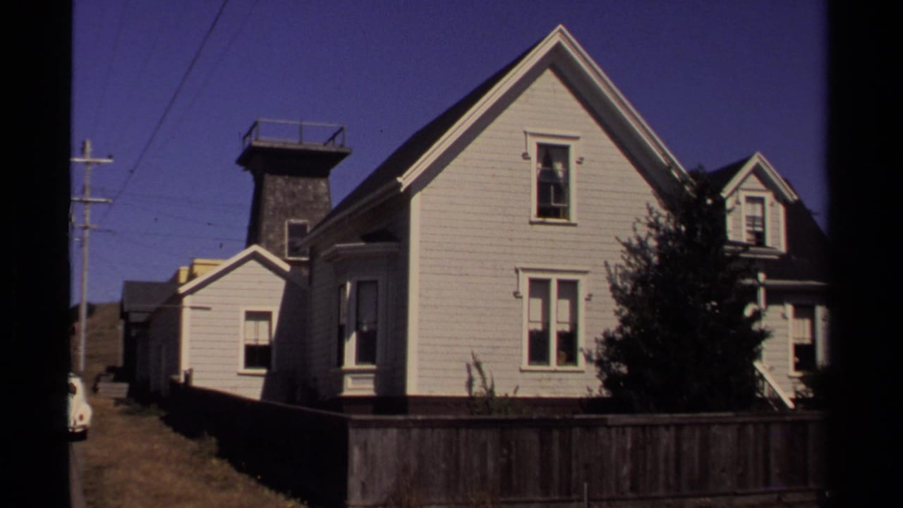 White clapboard house beside a fenced yard, with another structure and utility poles along the street.