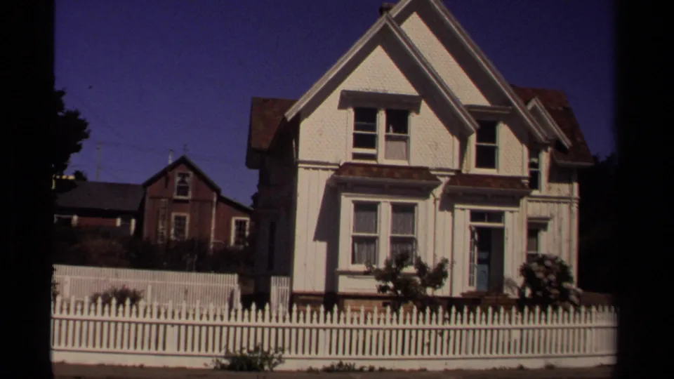 1980 Mendocino home-movie frame of a white residential house front, picket fence, shrubs, and background houses under blue sky.