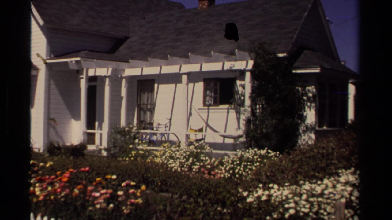 1980 8mm frame from Mendocino showing a white clapboard house, porch, dark roof, flowers, and garden shrubs in sunlight.