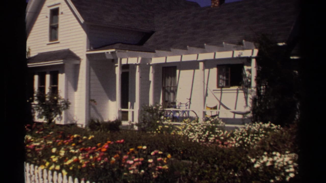 Sunlit white house facade with porch and dense flowers in the front yard.