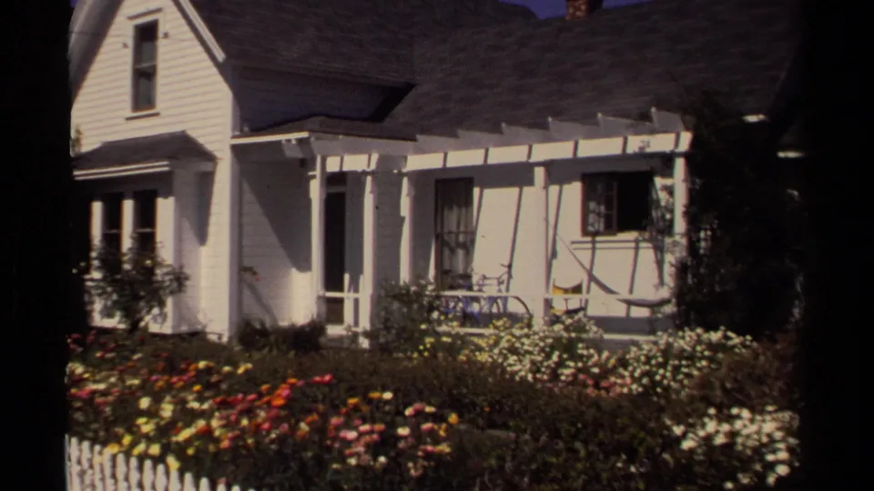 Mendocino residential exterior on 8mm film, with a white facade, porch, roofline, flower beds, and shrubs visible from the garden.