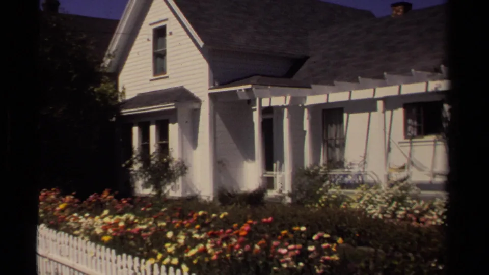 Angled 1980 Mendocino home-movie view of a white clapboard house with porch, picket fence, and red, yellow, and white garden blooms.