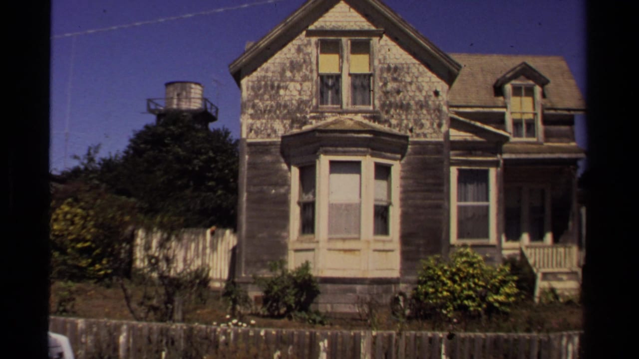 1980 8mm view of a weathered Victorian-style house in Mendocino, California, with peeling siding, bay window, porch, fence, and overgrown yard.