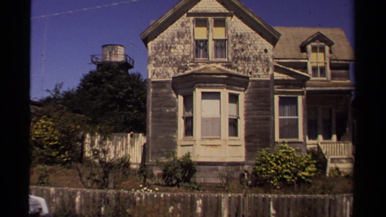 1980 8mm frame from Mendocino showing the same weathered house facade with bay window, porch railings, fence, shrubs, and roof dormer.