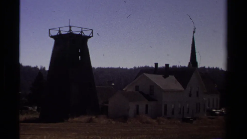 1980 8mm Mendocino view of a dark water tower silhouette near a white church-like building, pointed steeple, open grass, and distant tree line.