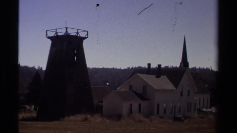 1980 8mm frame in Mendocino with a silhouetted water tower, white church-like building, steeple, rooflines, open ground, and distant wooded ridge.