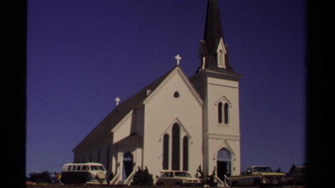 White church with steeple, crosses, and parked cars in bright sun.
