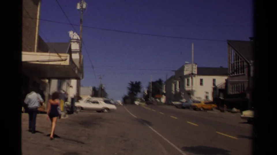 1980 Mendocino street scene with pedestrians walking past storefronts, parked cars, a yellow taxi-like car, and overhead utility wires.