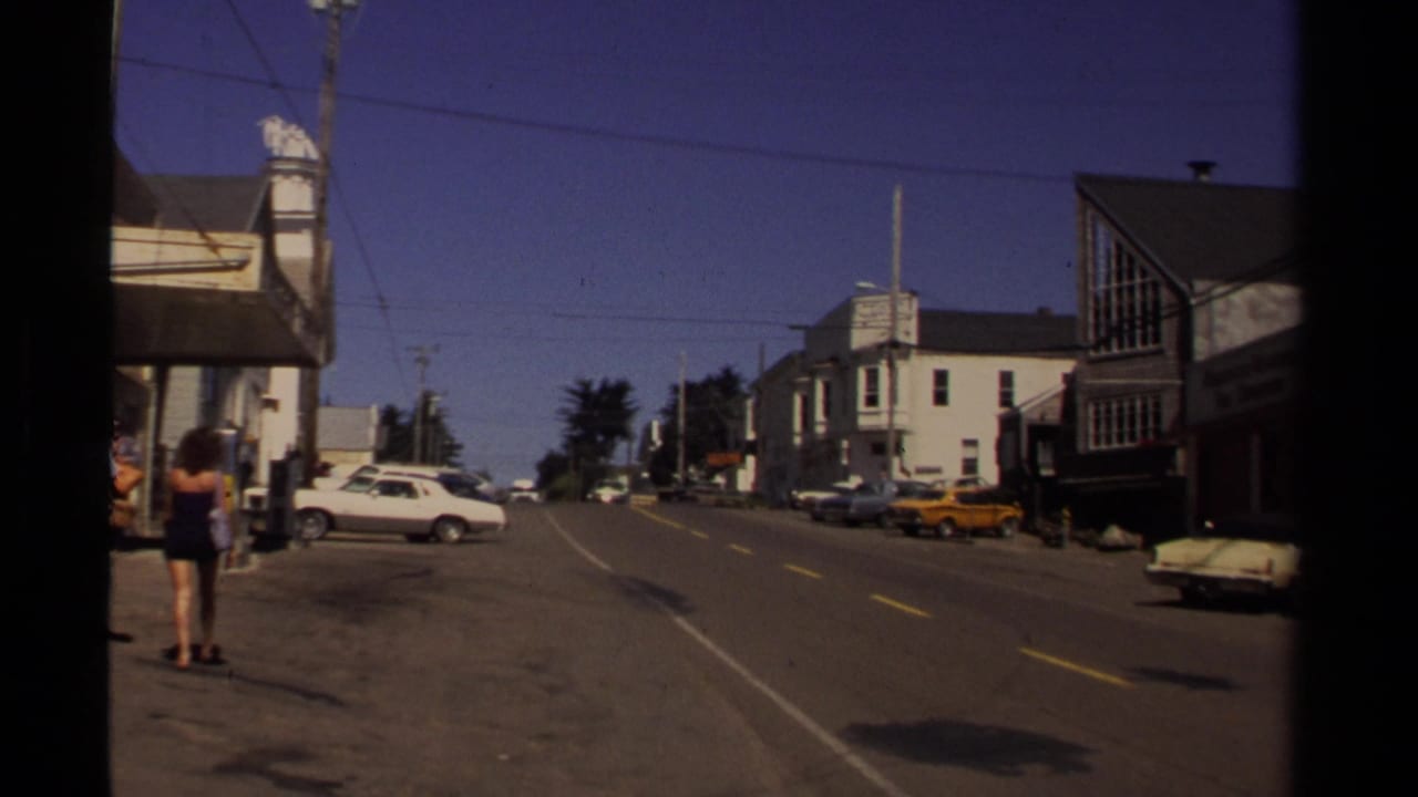 Sloping small-town street with parked cars, storefronts, utility lines, and pedestrians at the left edge.