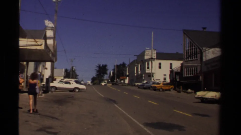 Mendocino, California street scene from supplied 1980 metadata, showing classic cars, storefronts, overhead wires, and pedestrians near the sidewalk.
