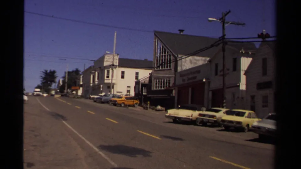Mendocino street corner from the 1980 clip, with parked period cars, storefront signage, utility pole, overhead wires, and sunlit pavement.