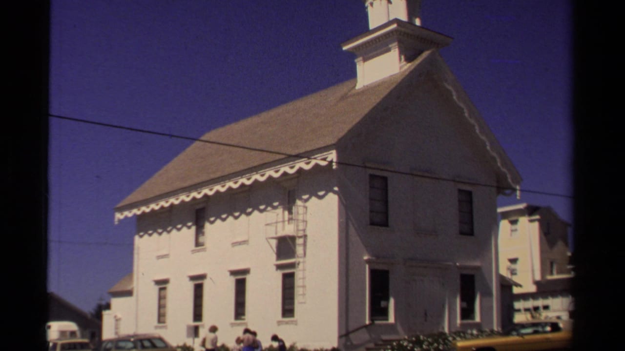 White church exterior with scalloped trim, parked cars, visitors, and blue sky.