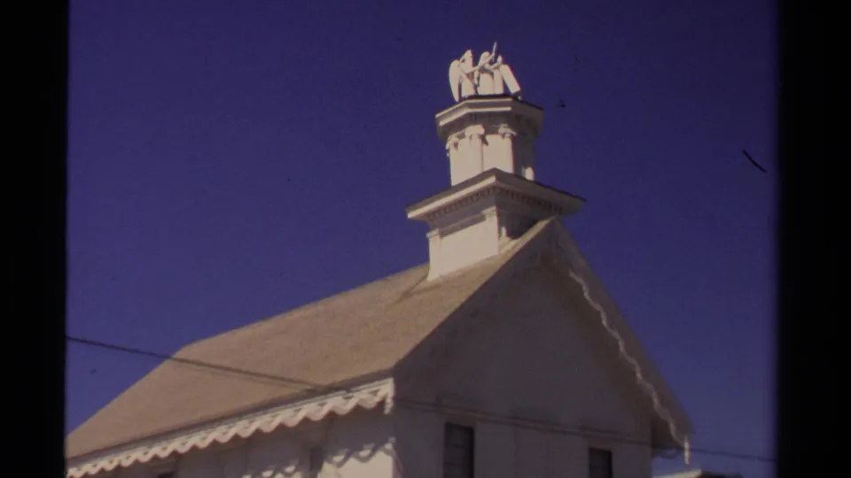 Upward 1980 Mendocino view of a white church roof, cupola, and angel statue on the steeple against a clear blue sky.