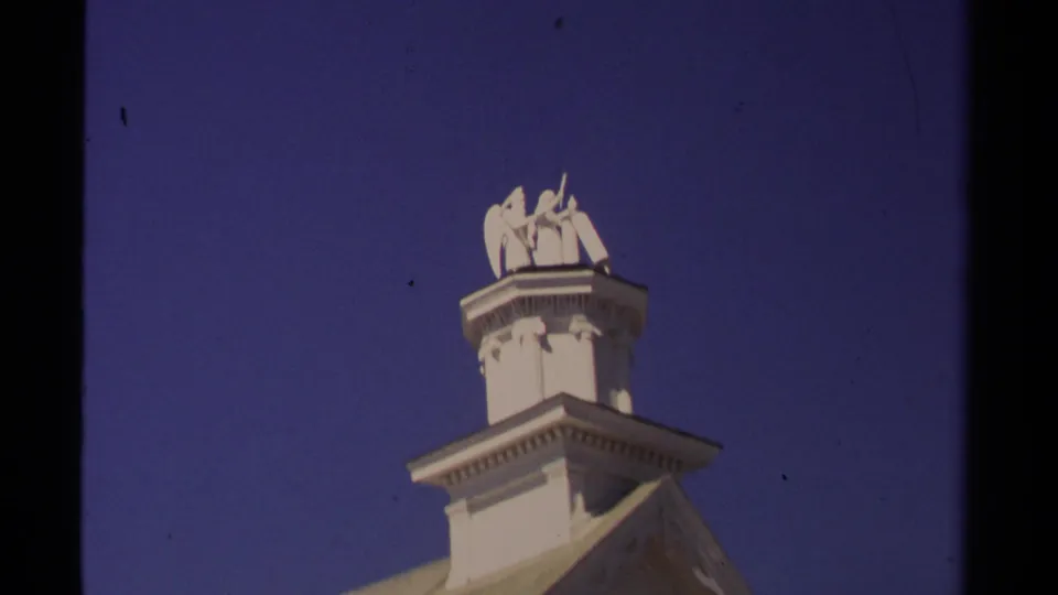 Close upward 1980 Mendocino shot of the white church cupola and angel statue, with the roof edge low in frame and blue sky behind.