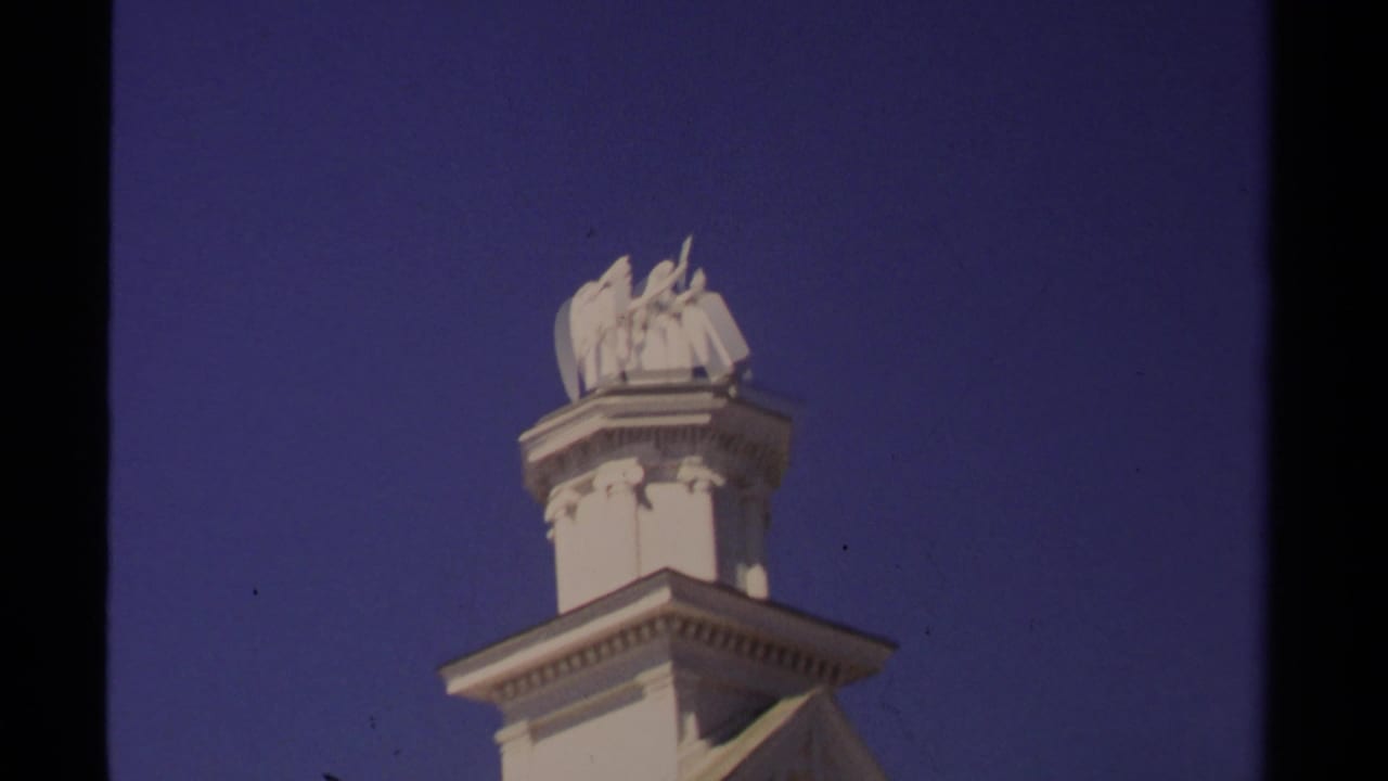 1980 Mendocino 8mm frame showing a white classical rooftop statue above a decorative building top against blue sky.