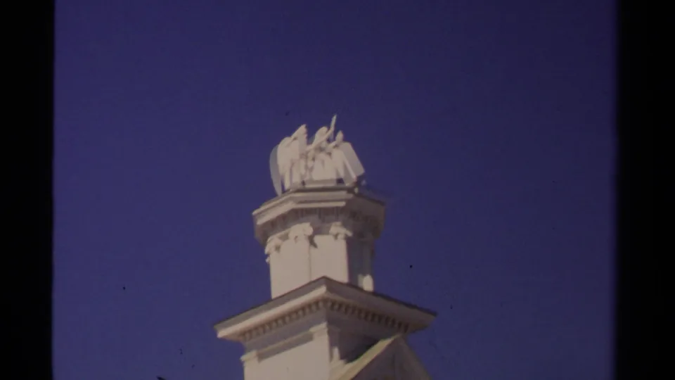 1980 Mendocino 8mm frame showing a white classical rooftop statue above a decorative building top against blue sky.