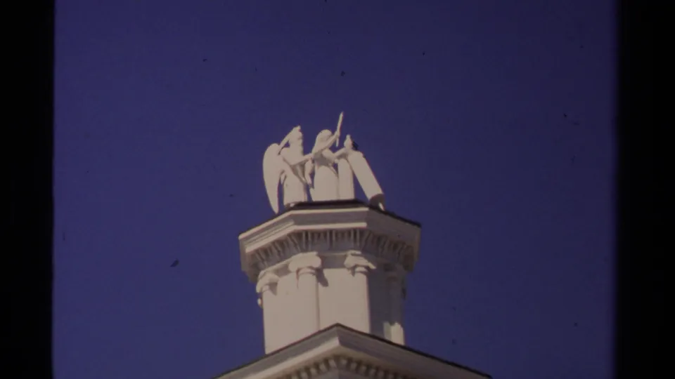 1980 Mendocino 8mm frame with white classical figures on a rooftop pedestal, seen from below against a clear blue sky.