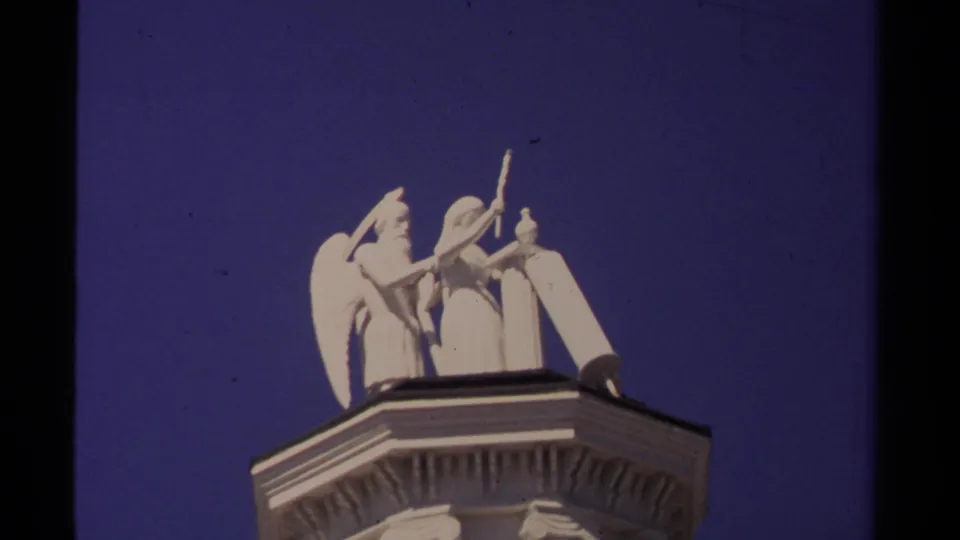 1980 Mendocino 8mm frame with the white rooftop statue group cropped above the building cornice under blue sky.